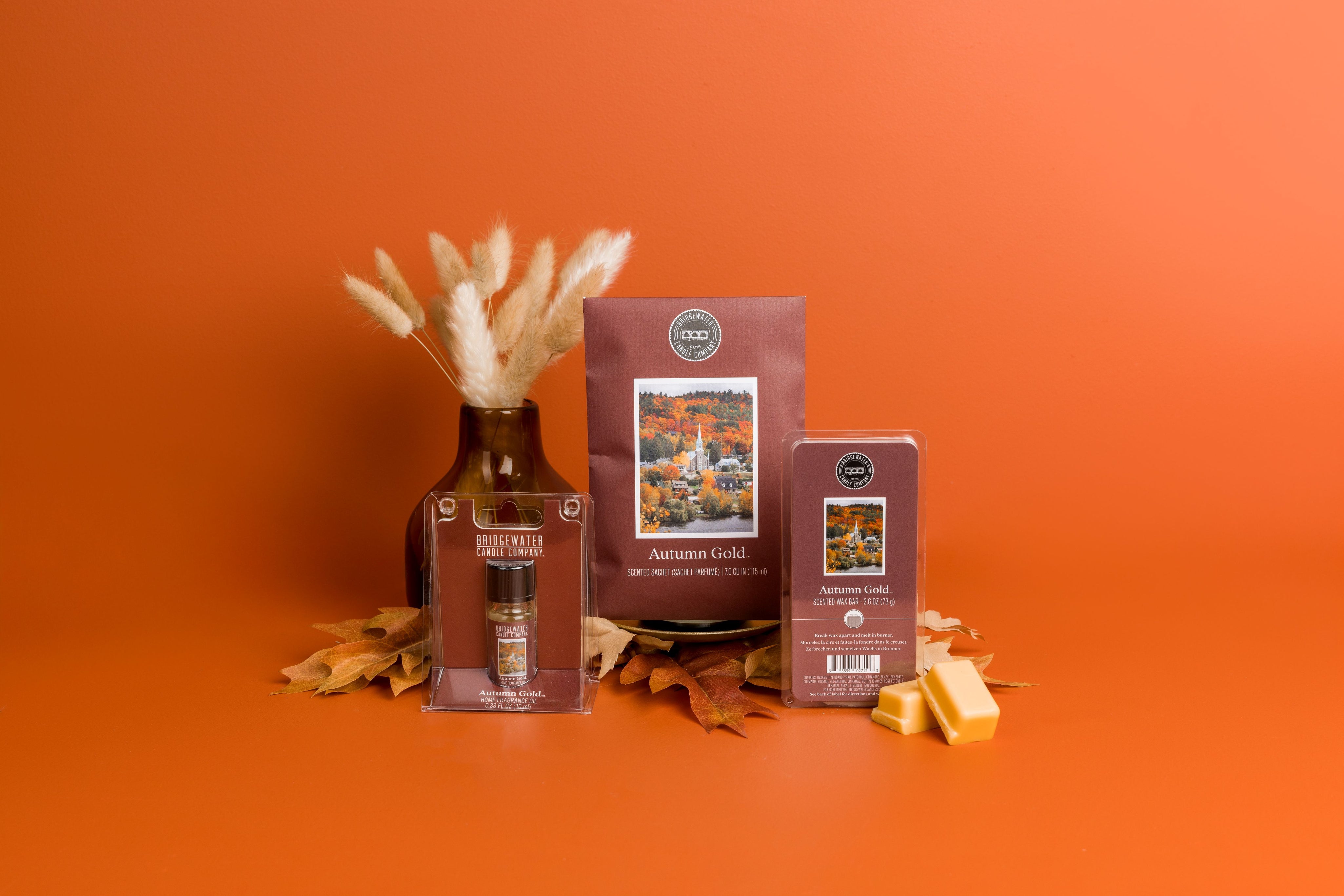 The Bridgewater Candle Autumn Gold Flameless Bundle, featuring a fragrance spray, soy-blend wax bar, and packet, is displayed on an orange surface with dried leaves and a vase of grasses against an orange backdrop.