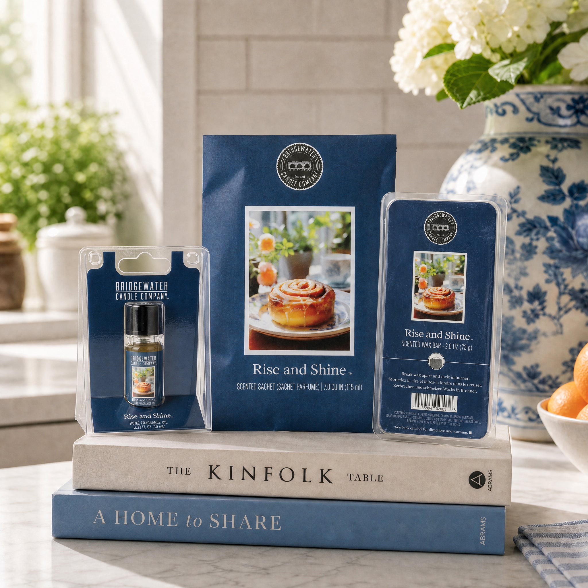 Bridgewater Candle's Rise & Shine Flameless Bundle rests atop stacked books near a vase of white flowers, while sunlight streams through a window in the background.