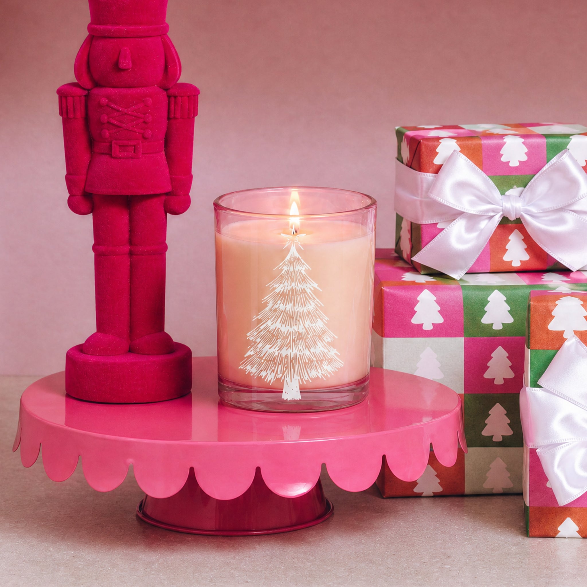 A pink nutcracker, a Sweet Grace Pink Tree Decal Jar Candle by Bridgewater Candle—featuring a festive pink soy-blend wax and silver tree design—and colorful Christmas gifts with white bows sit on a pink scalloped stand against a light pink background.