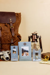 The Solitude Flameless Bundle by Bridgewater Candle features a brown backpack, vintage camera, lantern, compass, chocolate bar, scented sachet, and two landscape-cover blue booklets arranged on a beige background.