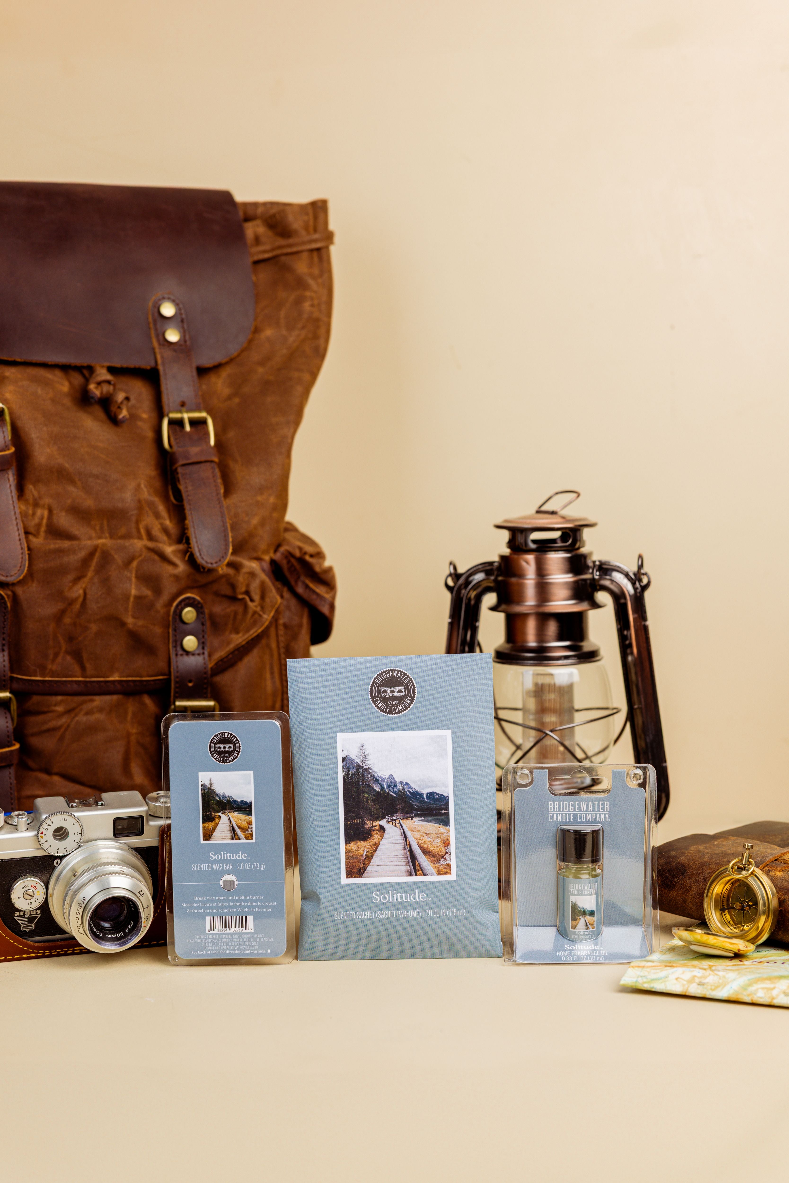 The Solitude Flameless Bundle by Bridgewater Candle features a brown backpack, vintage camera, lantern, compass, chocolate bar, scented sachet, and two landscape-cover blue booklets arranged on a beige background.