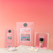 The Sunkissed Flameless Bundle from Bridgewater Candle, including a wax bar and home fragrance oil in beach-themed packaging, rests on sand with seashells against a pink gradient background, evoking a peaceful sunset.