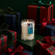 The I'll Be Home 8oz Jar Candle by Bridgewater Candle glows in a glass jar, surrounded by dark green, red, and plaid gift boxes with ribbon bows. A dark backdrop enhances the festive holiday mood and inviting Bridgewater fragrance.
