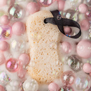 The Sweet Grace Body Wash Sponge by Bridgewater Candle, with a black ribbon, rests on soft bubbles—ready to exfoliate and pamper your skin with its creamy lather for a soothing, festive bath experience.