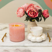 A Bridgewater Candle Sweet Grace Collection Candle #073, a white teacup on a pearlescent coaster, and pink peonies in a vase are arranged on a marble tray with gold handles atop a light-colored surface.