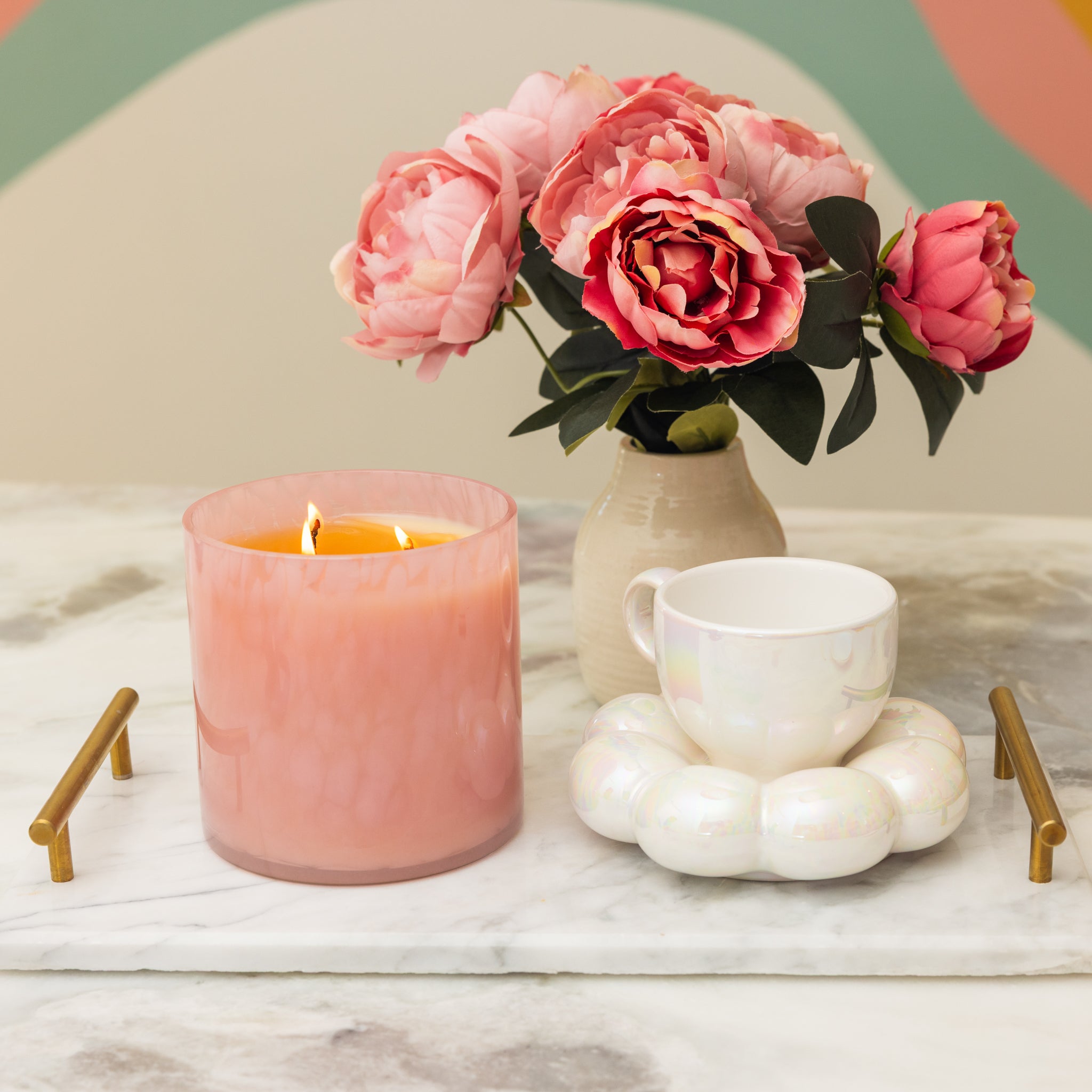A Bridgewater Candle Sweet Grace Collection Candle #073, a white teacup on a pearlescent coaster, and pink peonies in a vase are arranged on a marble tray with gold handles atop a light-colored surface.