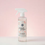 The Bridgewater Candle Sweet Grace Stain Remover is a vegan cleaning spray in a clear bottle with a white trigger nozzle, featuring pink and gray circular label design and displayed against a pink gradient background.