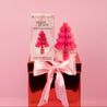 The Bridgewater Candle Sweet Grace Holiday Honeycomb Diffuser, in a pink honeycomb tree-shaped glass jar, sits on a red gift box with a pink bow. Its product box is behind it, all set against a solid pink background for festive fragrance display.