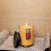 The Bridgewater Candle Autumn Gold Small Jar Candle, filled with soy-blend wax and topped with a black lid, glows in its glass jar on a patterned tray against a textured beige background.