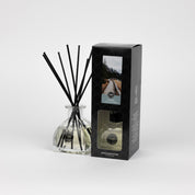 A Solitude Reed Diffuser by Bridgewater Candle with fiber reeds stands beside its black box, both featuring mountain landscape and text. These premium home fragrance items are displayed against a plain white background.