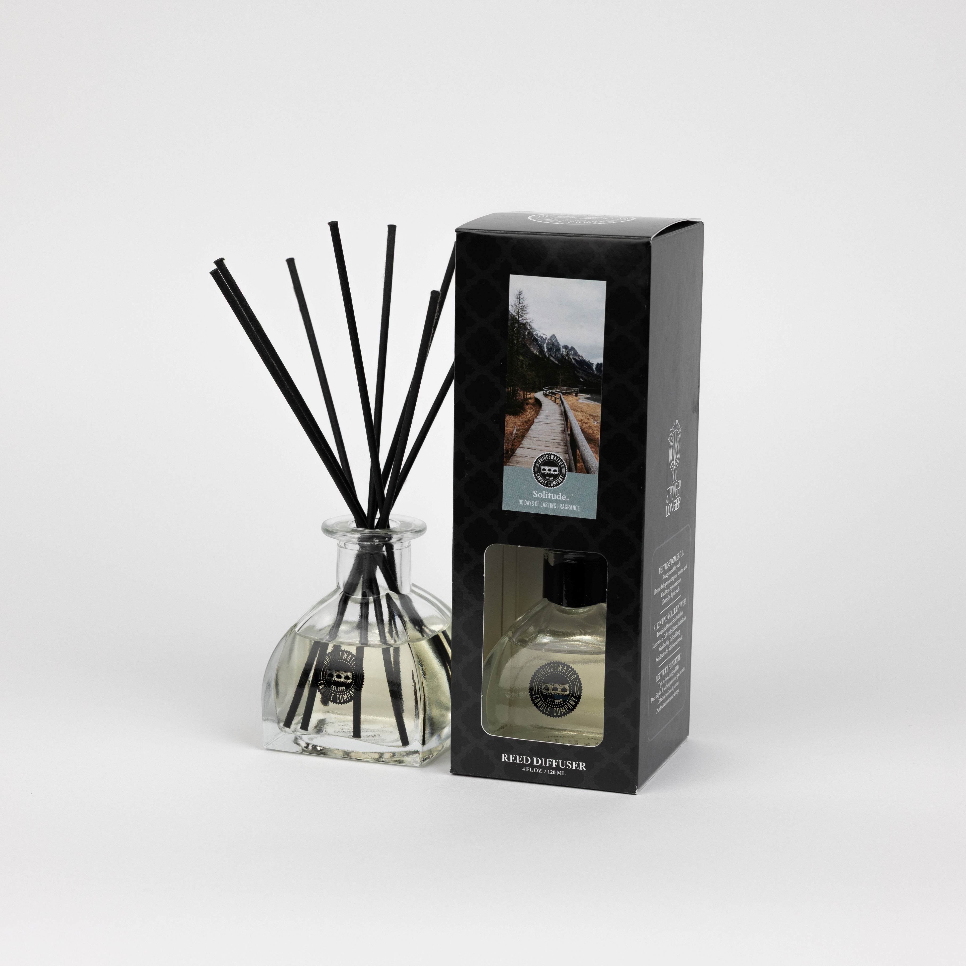 A Solitude Reed Diffuser by Bridgewater Candle with fiber reeds stands beside its black box, both featuring mountain landscape and text. These premium home fragrance items are displayed against a plain white background.