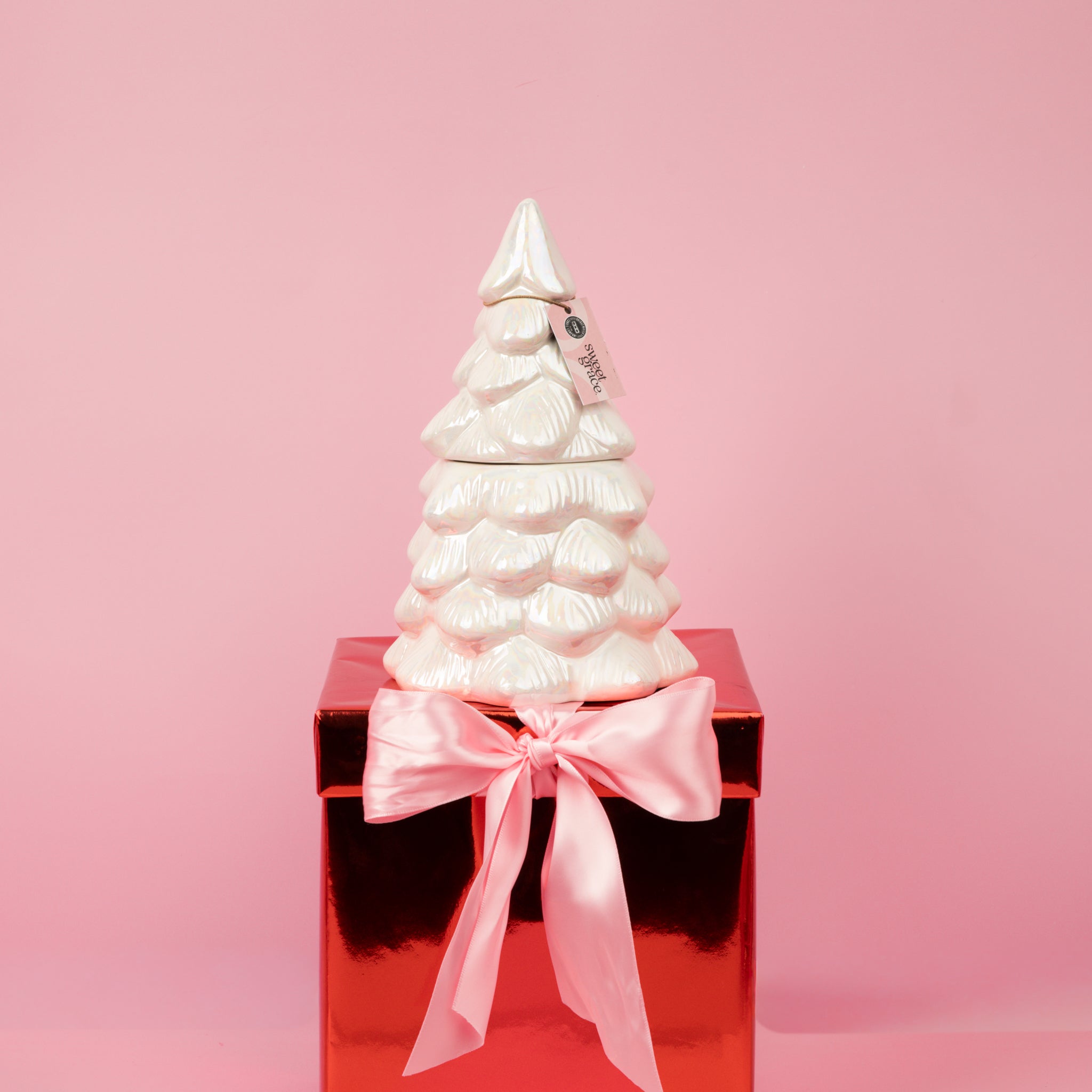 The Sweet Grace Collection Candle #70 by Bridgewater Candle sits atop a shiny red gift box with a large pink ribbon and bow, set against a pink background.