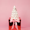 The Sweet Grace Collection Candle #70 by Bridgewater Candle sits atop a shiny red gift box with a large pink ribbon and bow, set against a pink background.