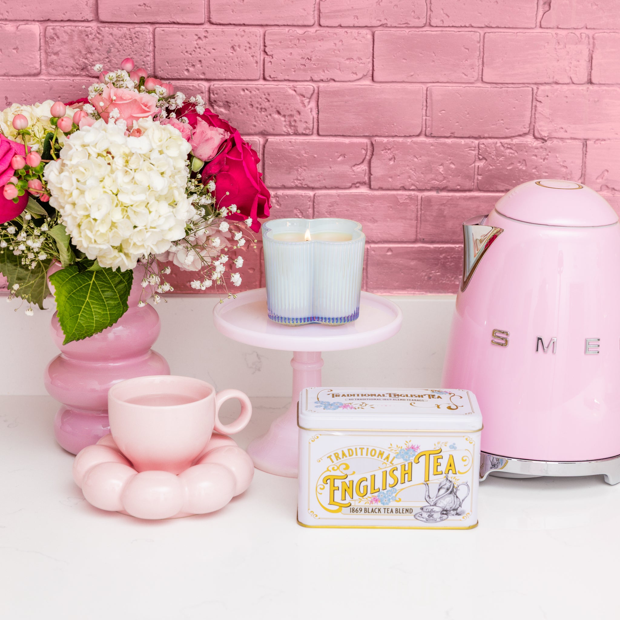 A pink SMEG electric kettle, traditional English tea in a tin, a pink cup and saucer, two Bridgewater Candle Sweet Grace Collection #077 pastel soy-blend candles, and a bouquet of pink and white flowers are set on a white surface against a pink brick wall.