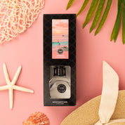 A boxed Bridgewater Candle Sunkissed Reed Diffuser with a coastal sunset design rests on a pink background, surrounded by a starfish, fiber reeds, a straw hat with beige ribbon, netting, and green palm leaves.