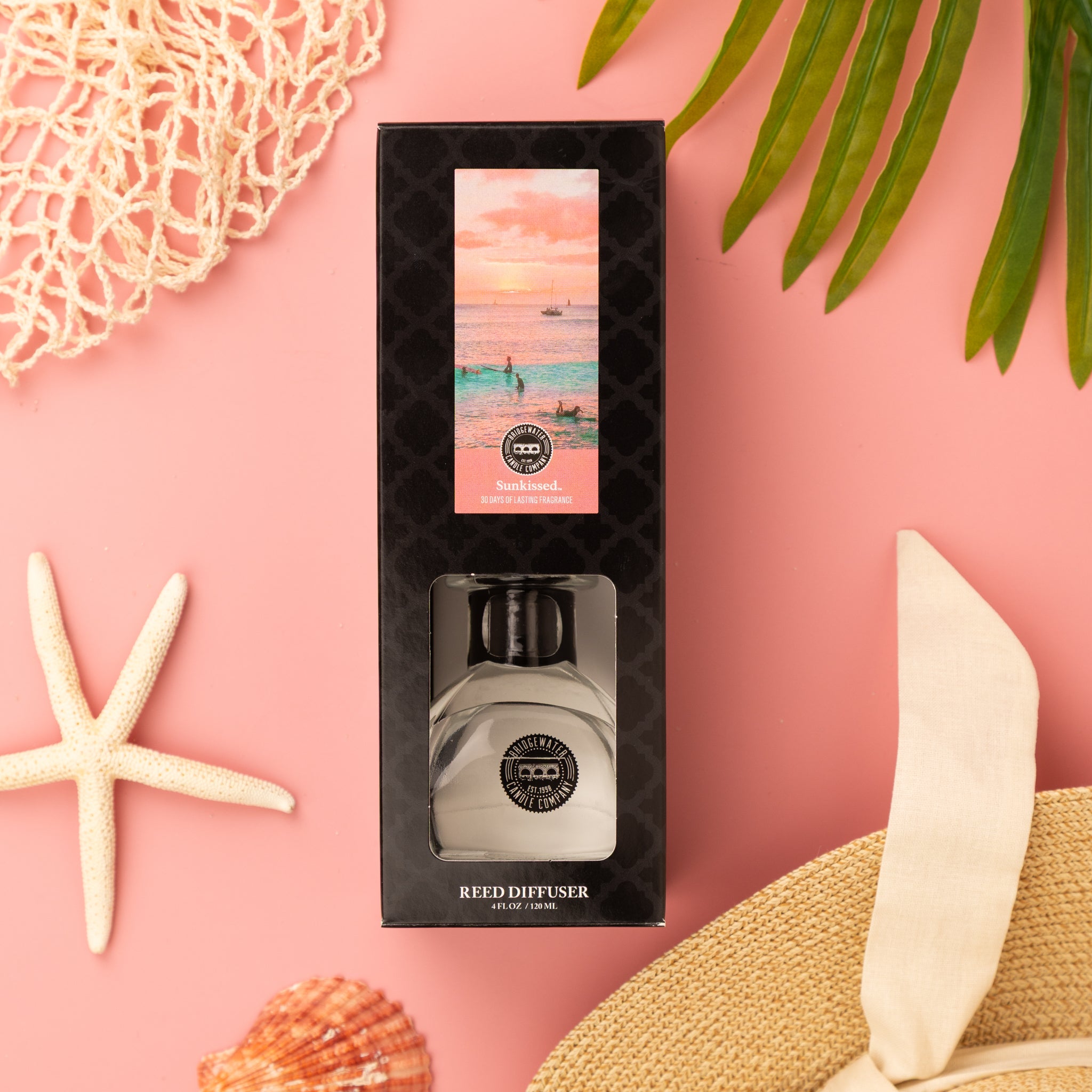 A boxed Bridgewater Candle Sunkissed Reed Diffuser with a coastal sunset design rests on a pink background, surrounded by a starfish, fiber reeds, a straw hat with beige ribbon, netting, and green palm leaves.