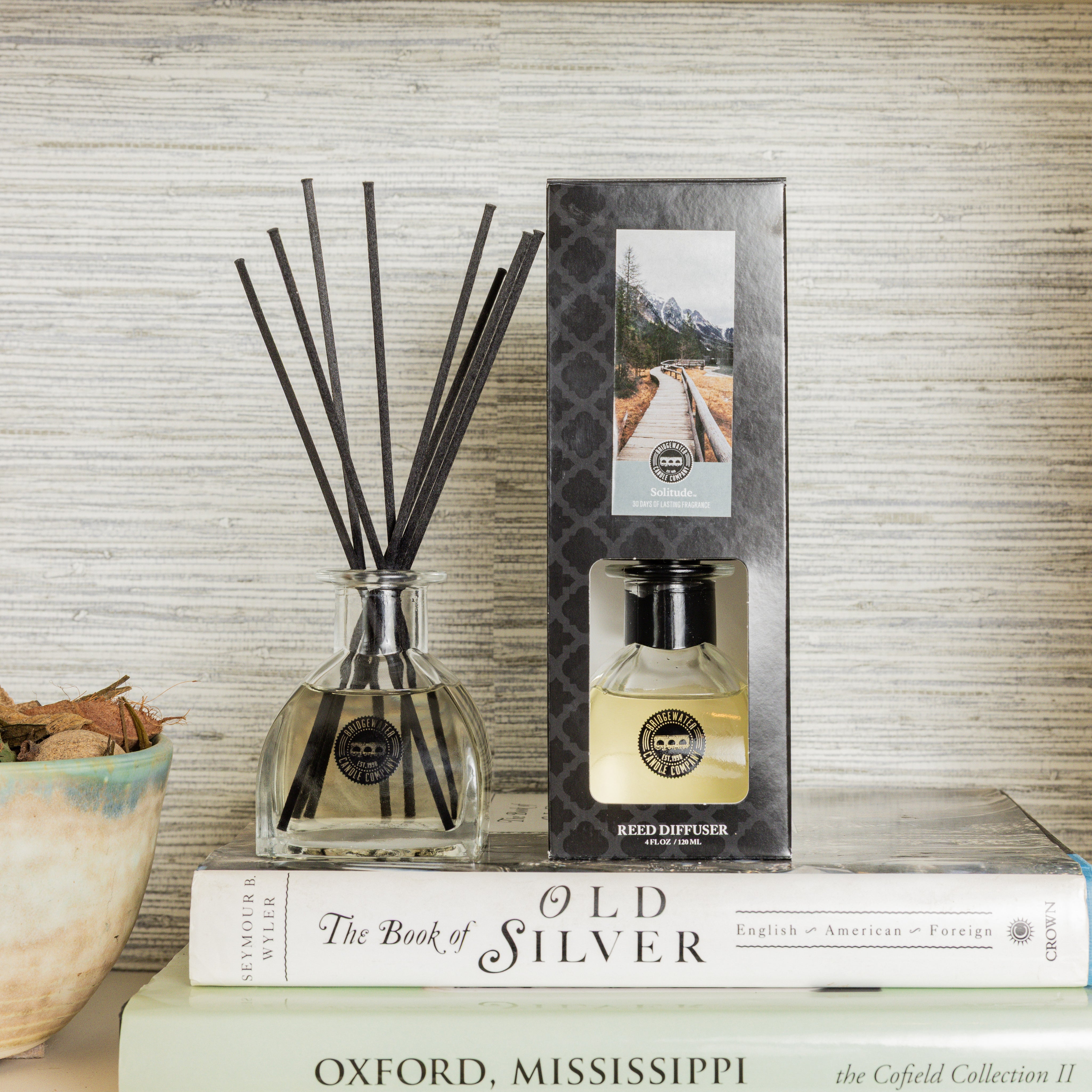A Bridgewater Candle Solitude Reed Diffuser with fiber reeds and its box sits atop stacked books, including The Book of Old Silver and Oxford, Mississippi. Dried leaves in a small bowl enhance the premium home fragrance atmosphere.
