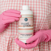 Wearing a pink checkered dress and pink gloves, someone holds a bottle of Bridgewater Candle Sweet Grace Cleaning Concentrate | Mop Soap, ready to revitalize their space with this fragrant cleaner.