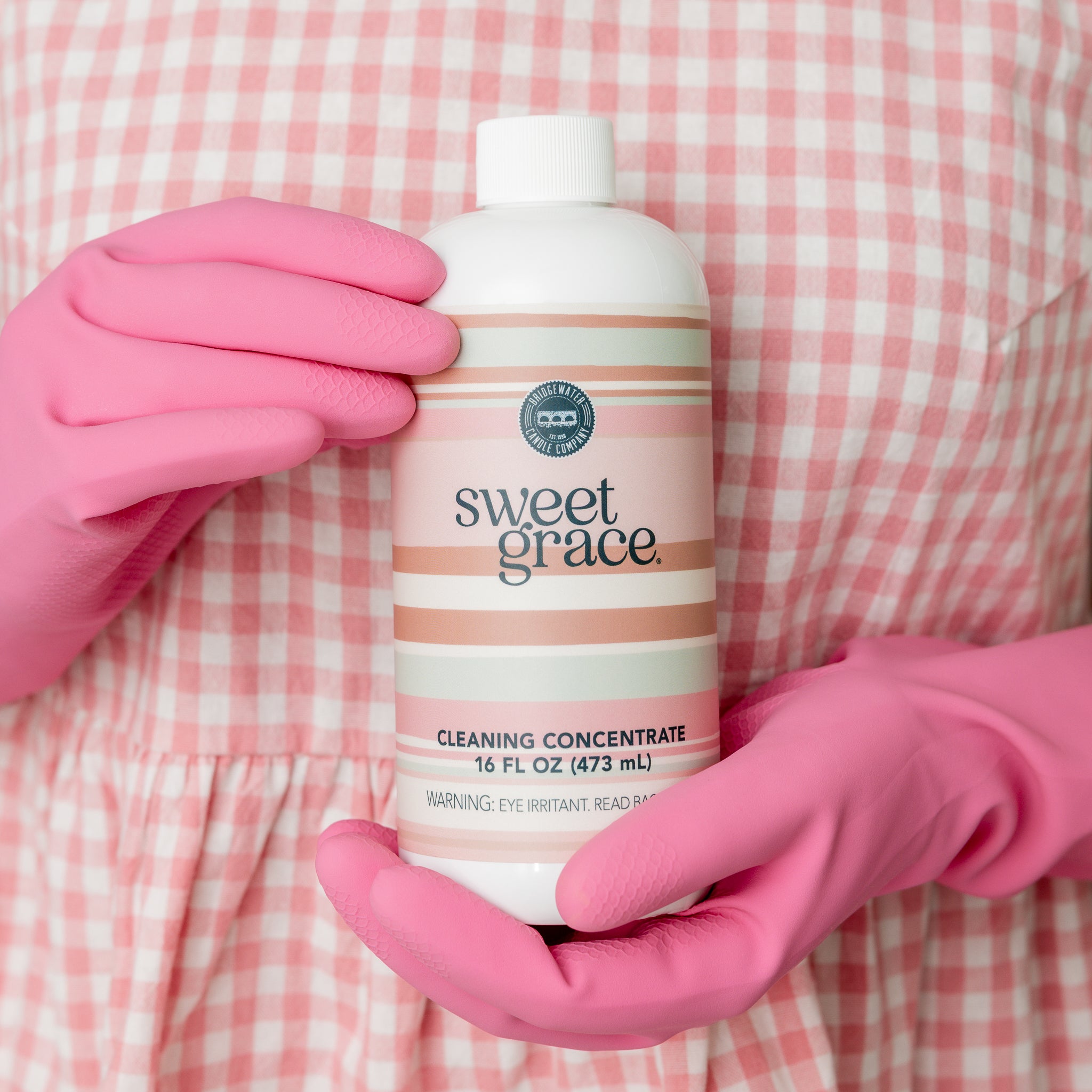 Wearing a pink checkered dress and pink gloves, someone holds a bottle of Bridgewater Candle Sweet Grace Cleaning Concentrate | Mop Soap, ready to revitalize their space with this fragrant cleaner.