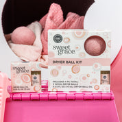 The Bridgewater Candle Sweet Grace Dryer Ball Kit, featuring wool dryer balls and fragranced laundry oil, is shown on a pink basket with soft laundry and a dryer ball nearby. The packaging displays pastel circles and product details.