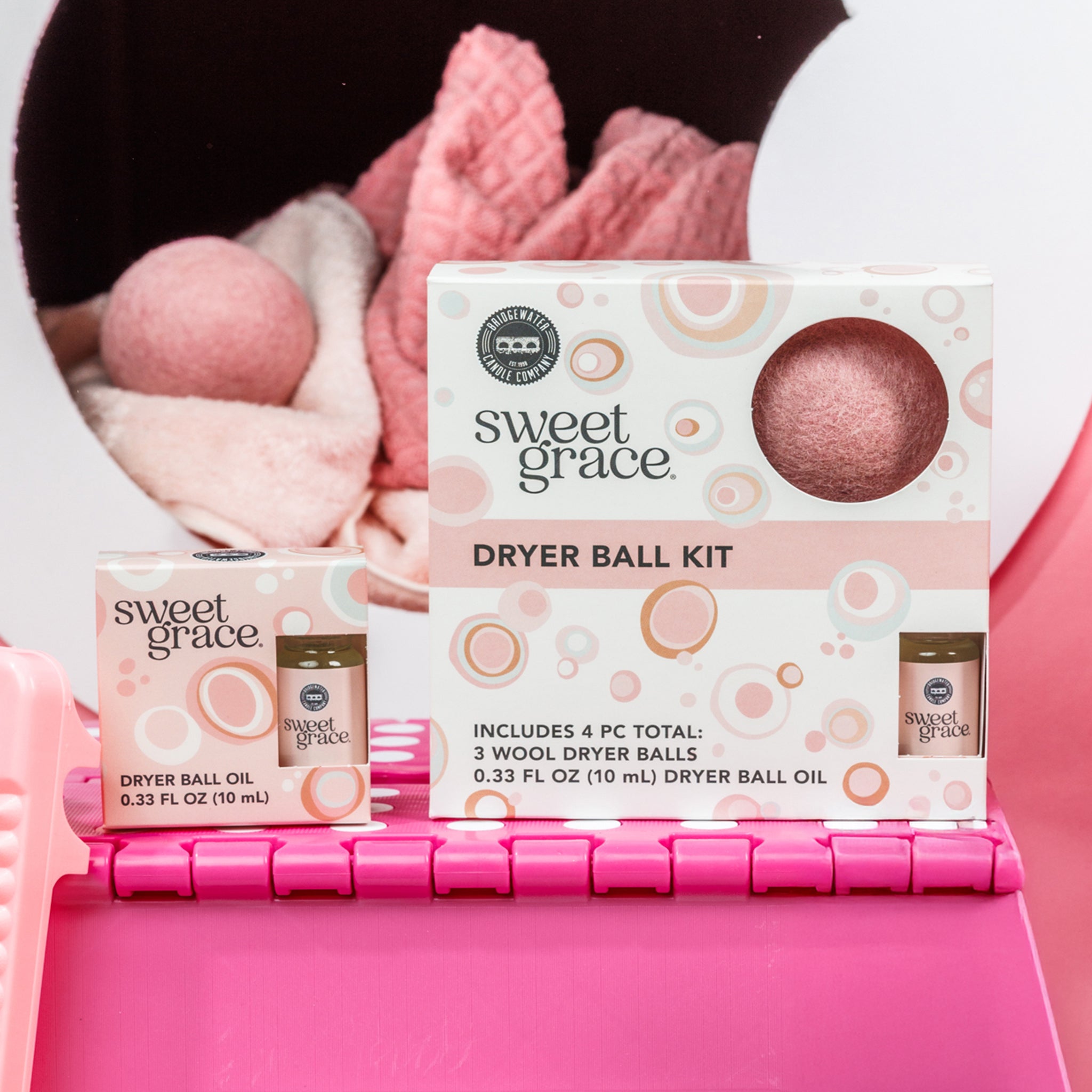 The Bridgewater Candle Sweet Grace Dryer Ball Kit, featuring wool dryer balls and fragranced laundry oil, is shown on a pink basket with soft laundry and a dryer ball nearby. The packaging displays pastel circles and product details.