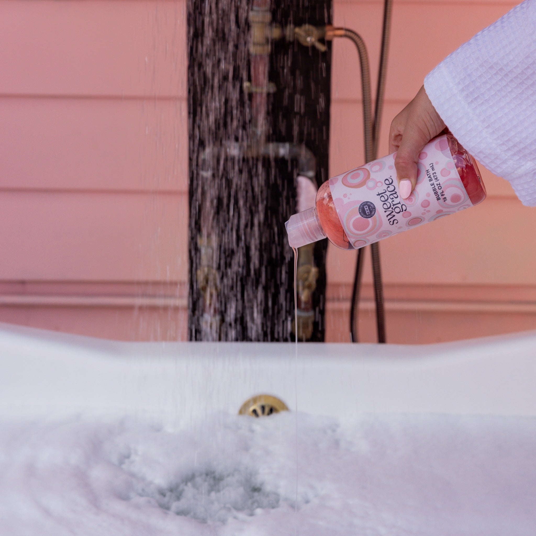 A hand in a white robe pours Sweet Grace Bubble Bath into a bathtub under a shower head.