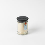 The High Bloom 8oz Small Jar Candle by Bridgewater Candle features soy-blend wax in a clear glass jar with a black lid and branded label. A colorful tag highlights the scent, set against a plain white background.