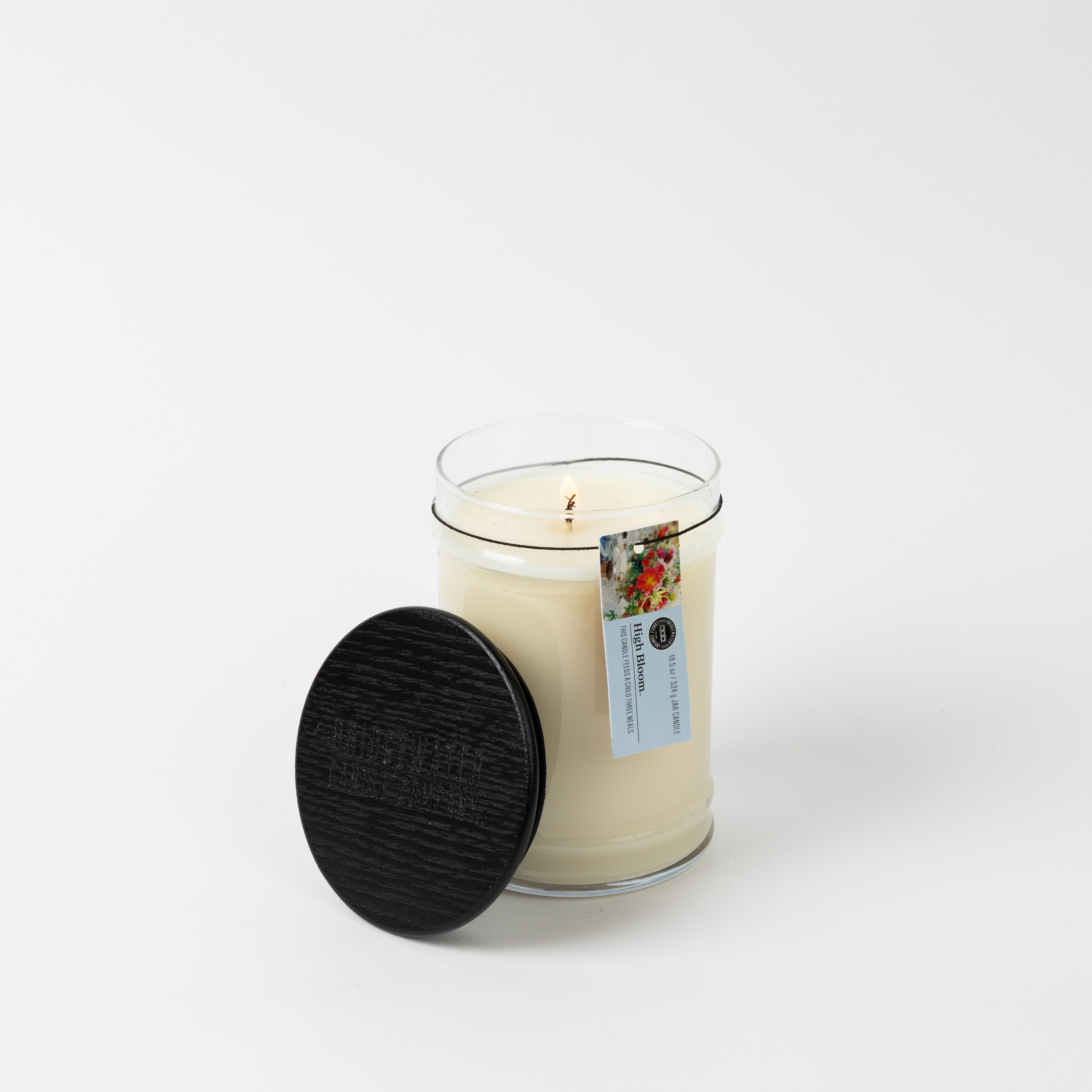 A lit High Bloom 18oz Large Jar Candle by Bridgewater Candle, made from soy-blend wax, sits in a clear glass jar with a dark wooden lid beside it. A small floral label accents the white candle against a plain white background.