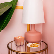A round glass table holds a Bridgewater Candle Sweet Grace Collection Candle #075 in a flower-shaped vessel, a small white plate with gold jewelry, and a pink lamp with a white shade, all framed by pink walls and a large green leaf.