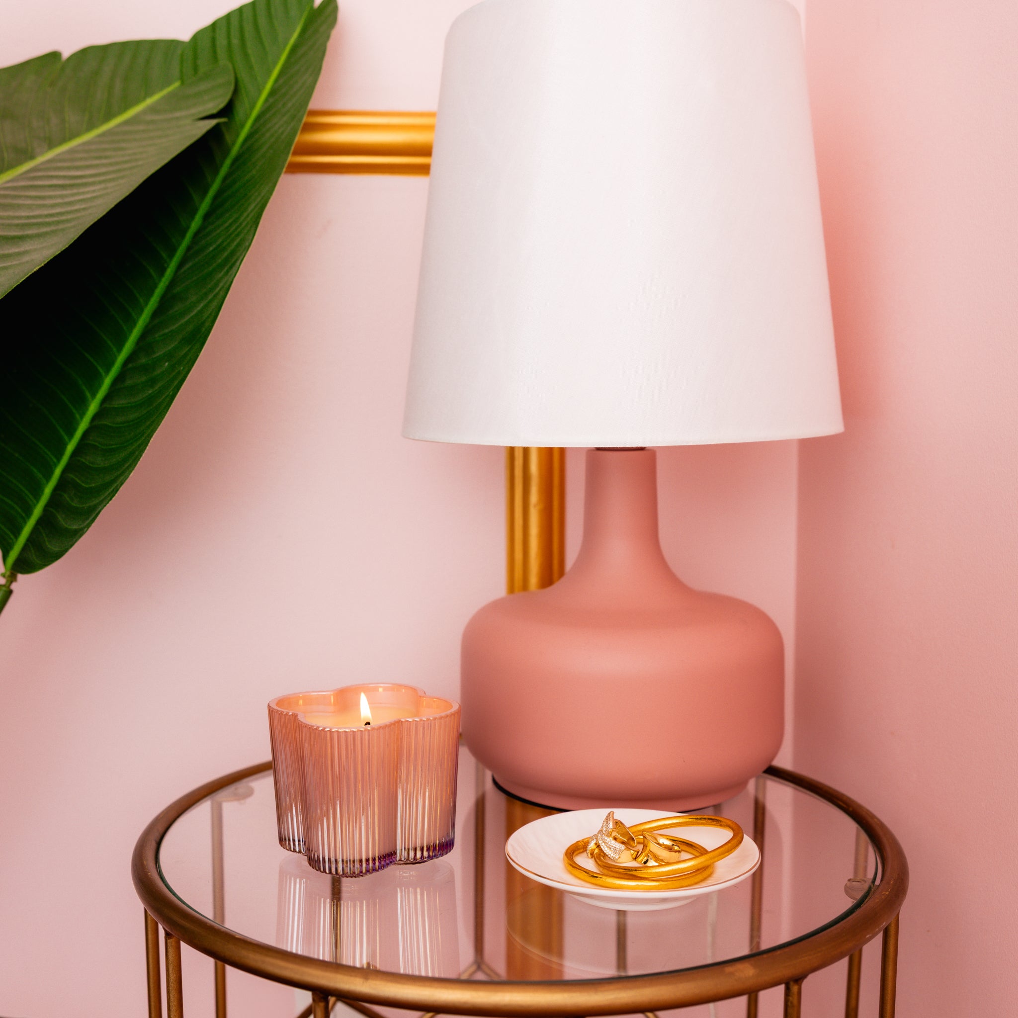 A round glass table holds a Bridgewater Candle Sweet Grace Collection Candle #075 in a flower-shaped vessel, a small white plate with gold jewelry, and a pink lamp with a white shade, all framed by pink walls and a large green leaf.