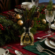 A holiday table features pinecones, gold ornaments, evergreen branches, plaid napkins, and glassware. At the center sits the Bridgewater Candle "I'll Be Home" Reed Diffuser with fiber reeds, filling the festive setting with cozy home fragrance.
