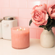 A Bridgewater Candle Sweet Grace Collection Candle #073 with pink soy-blend wax burns beside a white vase of pink peonies, against white canisters and a pink tiled wall.