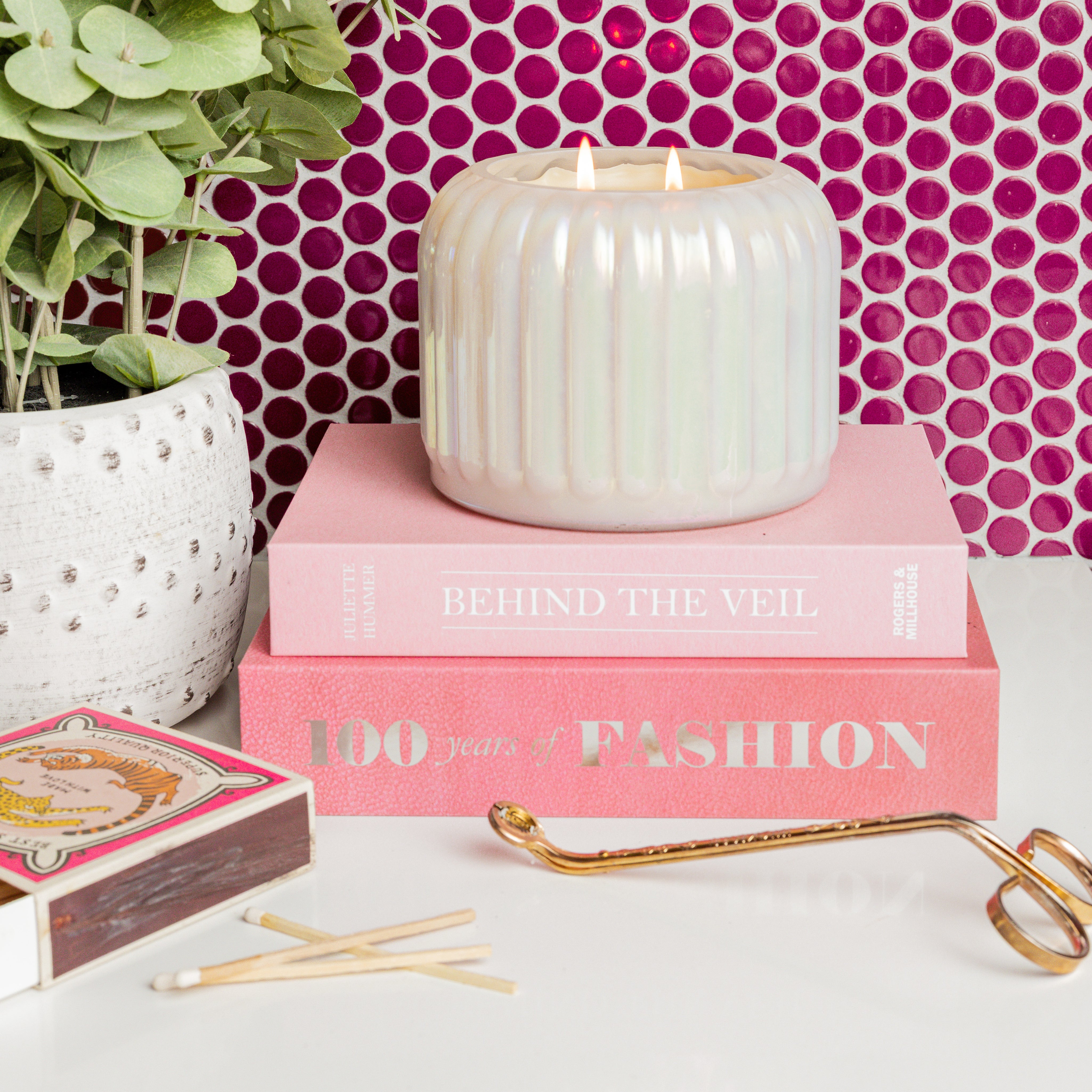 A Bridgewater Candle Sweet Grace Collection Candle #071 burns atop two pink books, 100 Years of Fashion and Behind the Veil, with a potted plant, matches, and a gold candle snuffer nearby on a white surface.