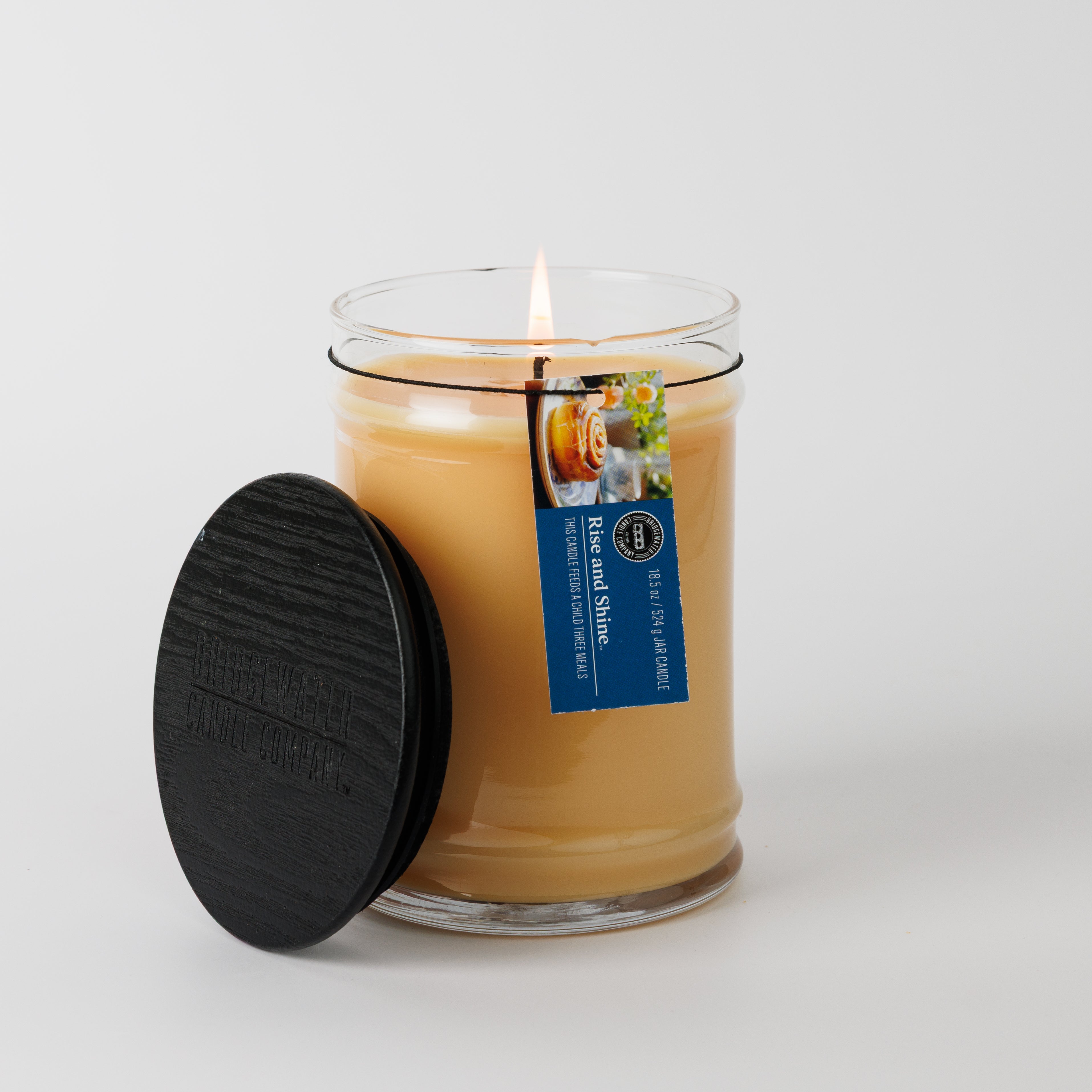 A lit Bridgewater Candle Rise & Shine 18oz Large Jar Candle sits in a clear glass jar with a black lid nearby. Made with soy-blend wax, it features a blue tag labeled Renew and Shine, plus a chamomile flower image. The background is white.