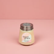 The Bridgewater Candle Sweet Grace Collection Candle #078, featuring cream-colored soy-blend wax in a petite glass jar with a metal lid, sits on a pink surface with a pastel label against a matching background.