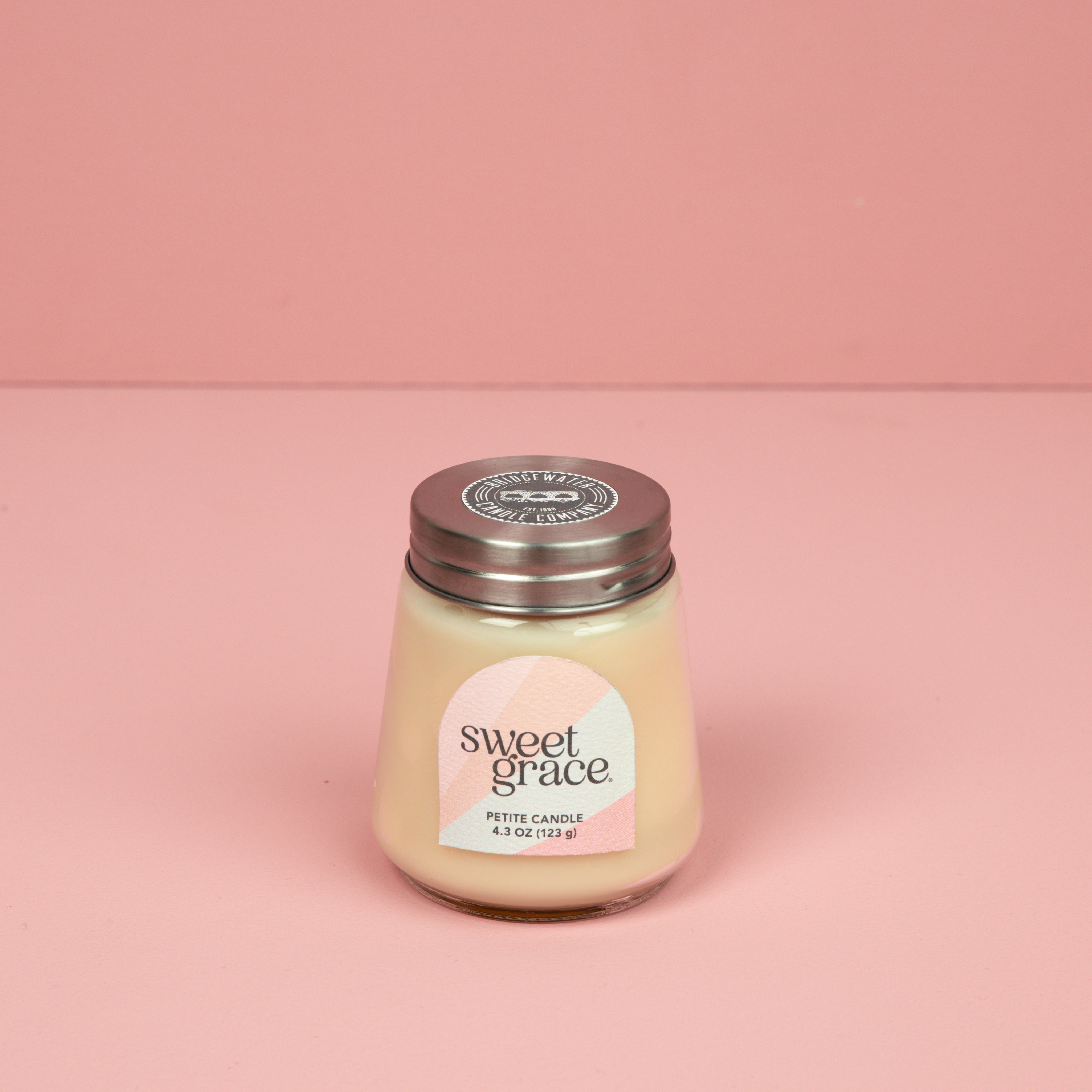 The Bridgewater Candle Sweet Grace Collection Candle #078, featuring cream-colored soy-blend wax in a petite glass jar with a metal lid, sits on a pink surface with a pastel label against a matching background.