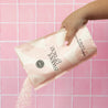 A hand pours Bridgewater Candle’s Sweet Grace Soaking Salts, enriched with Pink Himalayan Sea Salt, from a pink and cream bag against a pink tiled background.