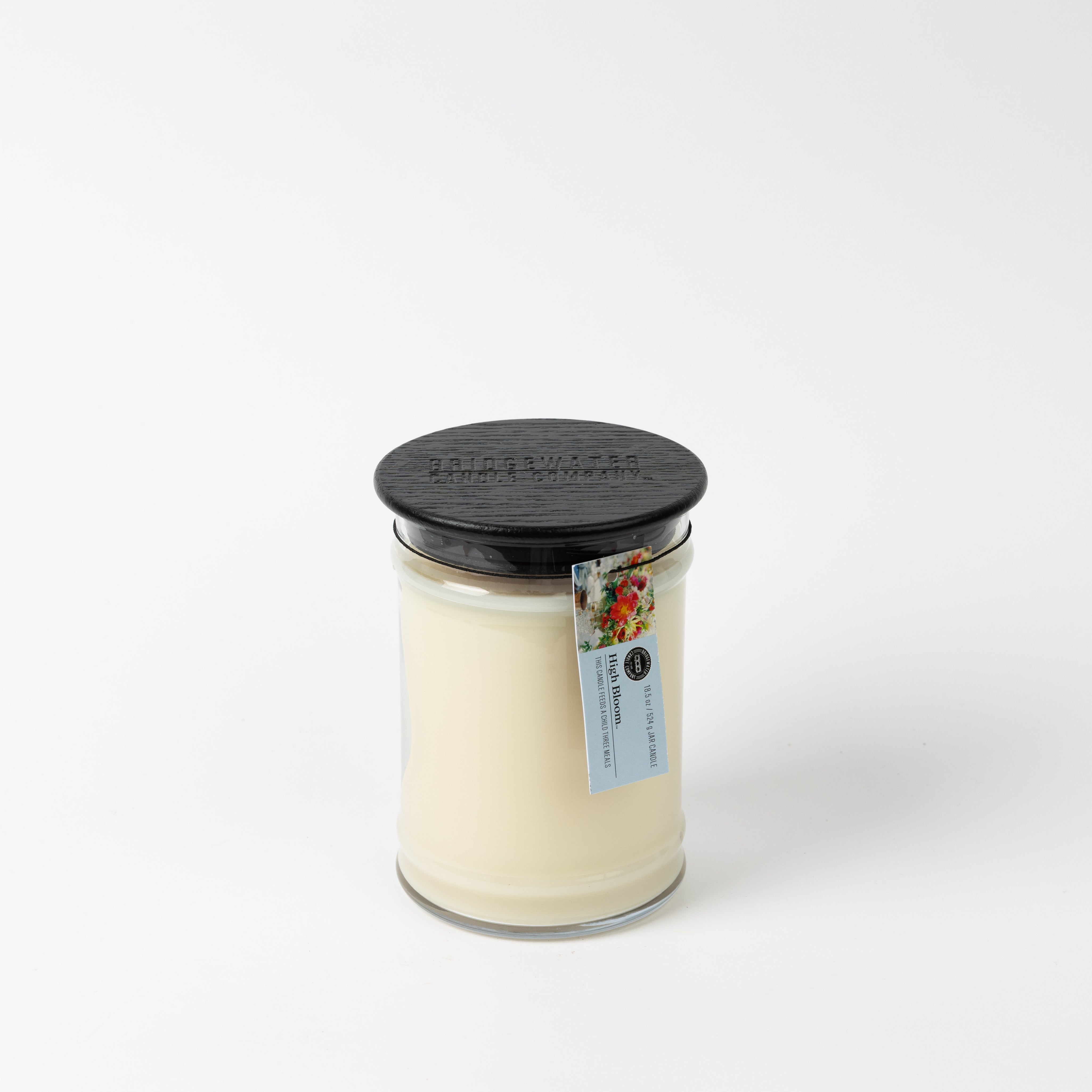 The High Bloom 18oz Large Jar Candle by Bridgewater Candle features creamy soy-blend wax, a black textured lid, and a floral tag, set against a white background. Enjoy its inviting fragrance in any room.