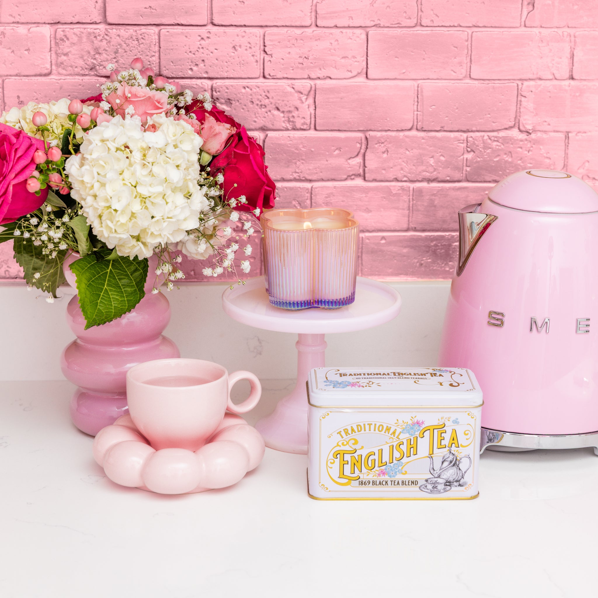 A pastel pink kitchen with a pink kettle, cup and saucer, a bouquet of white and pink flowers in a flower-shaped vase, a Bridgewater Candle Sweet Grace Collection Candle #075 (soy-blend), and an English Tea tin against a pink brick wall.