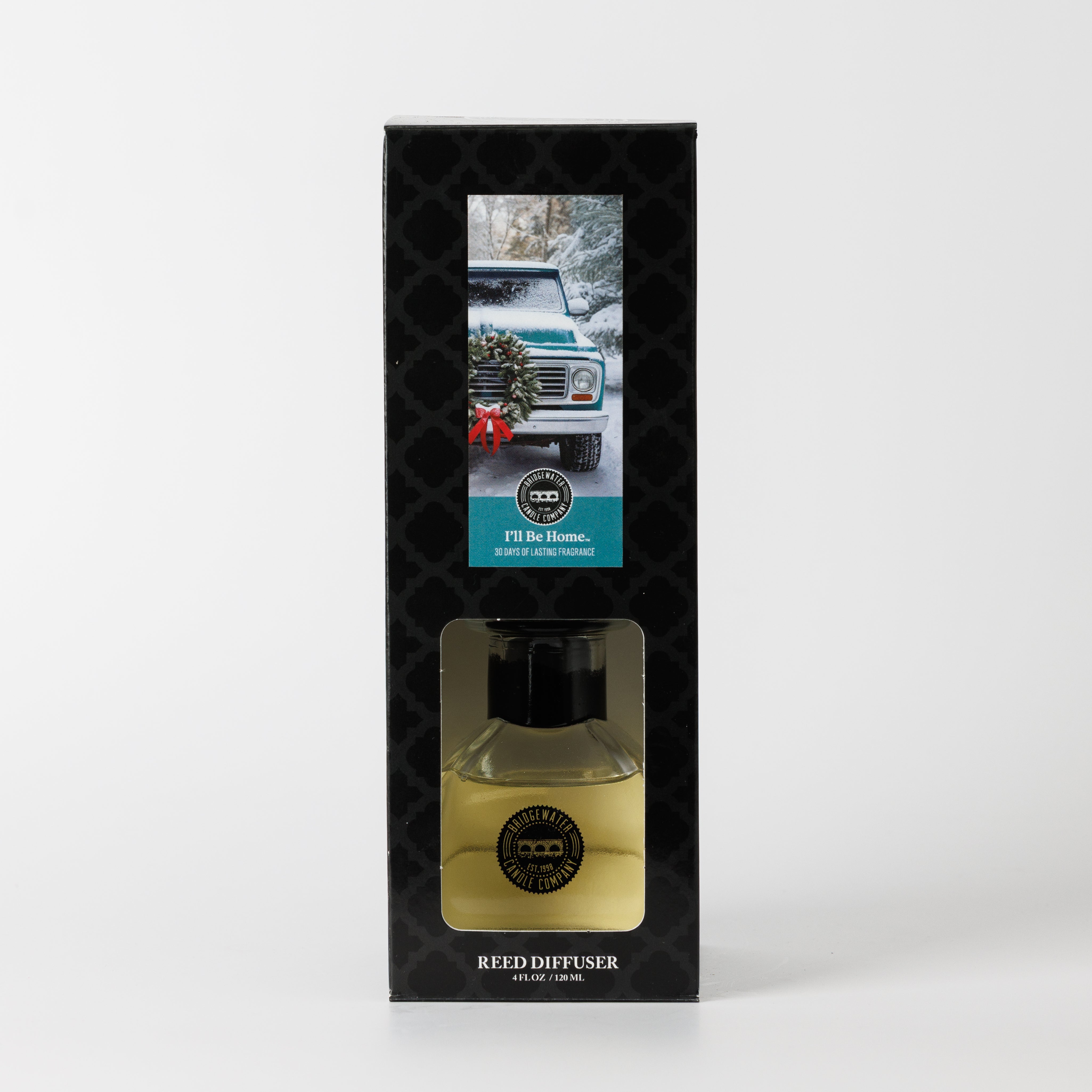 The Bridgewater Candle "I'll Be Home" Reed Diffuser boxed set features a winter scene with a white SUV and wreath. The clean home fragrance, diffuser bottle, and fiber reeds are visible through the packaging.