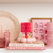 A shelf displays pink glassware, a pink plate, books, a Bridgewater Candle Sweet Grace Honeycomb Diffuser on stacked books, and a framed illustration of coffee makers, all styled in a cohesive pink color scheme.