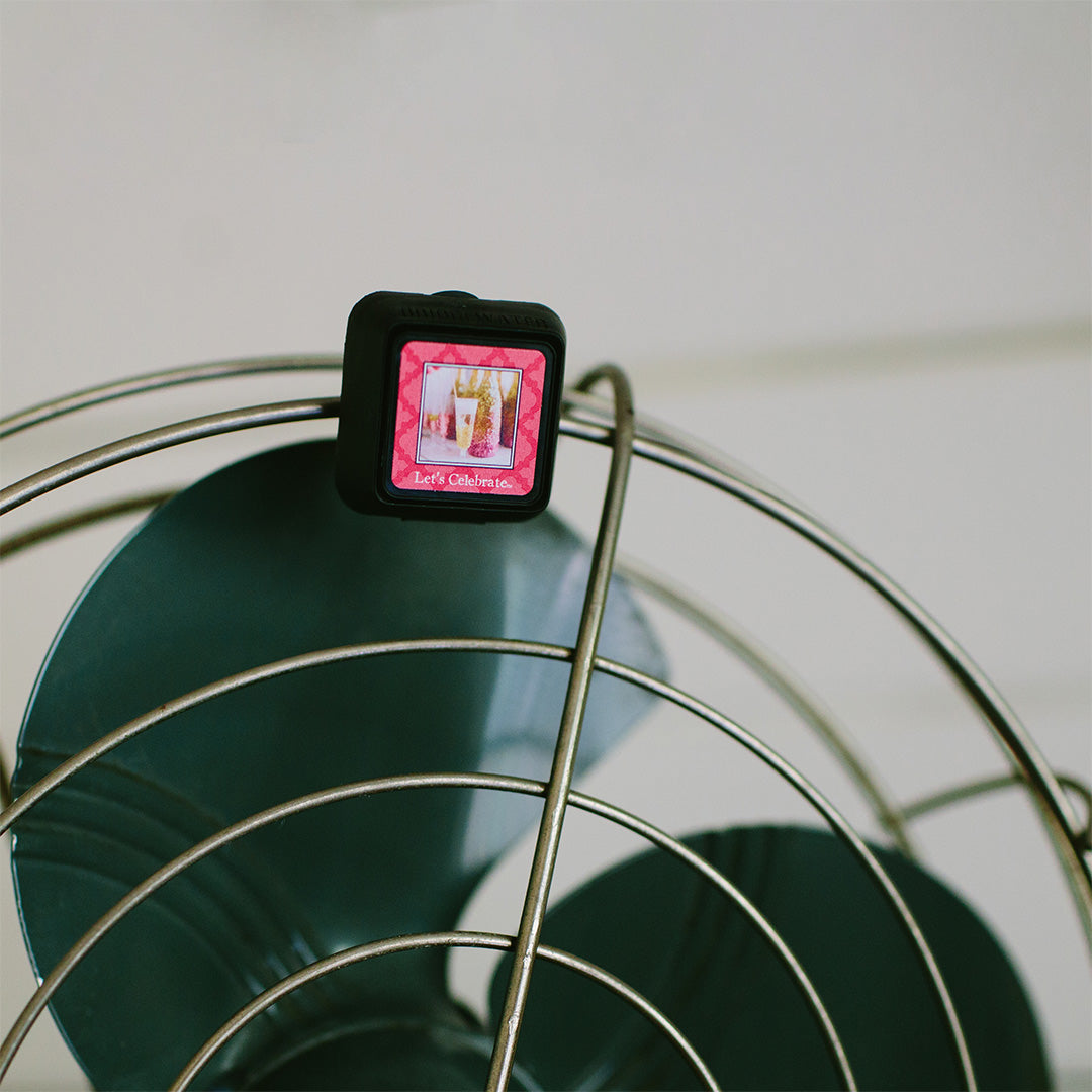 A square Auto Vent Clip-Let’s Celebrate air freshener by Bridgewater Candle clipped to the metal wire cage of a fan, featuring a pink graphic that reads Lets Celebrate.