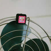 A square Auto Vent Clip-Let’s Celebrate air freshener by Bridgewater Candle clipped to the metal wire cage of a fan, featuring a pink graphic that reads Lets Celebrate.