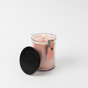 A lit Sunkissed 18oz Large Jar Candle by Bridgewater Candle, made with soy-blend wax, sits in a clear glass jar on a white background. Its black lid rests beside it and a small tag is attached to the jar.