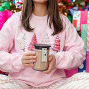 A person in pink and white pajamas and a pink Christmas tree sweatshirt holds a Bridgewater Candle Sweet Grace 18oz Large Jar Candle, with colorful gifts and a decorated tree in the background for festive fragrance charm.