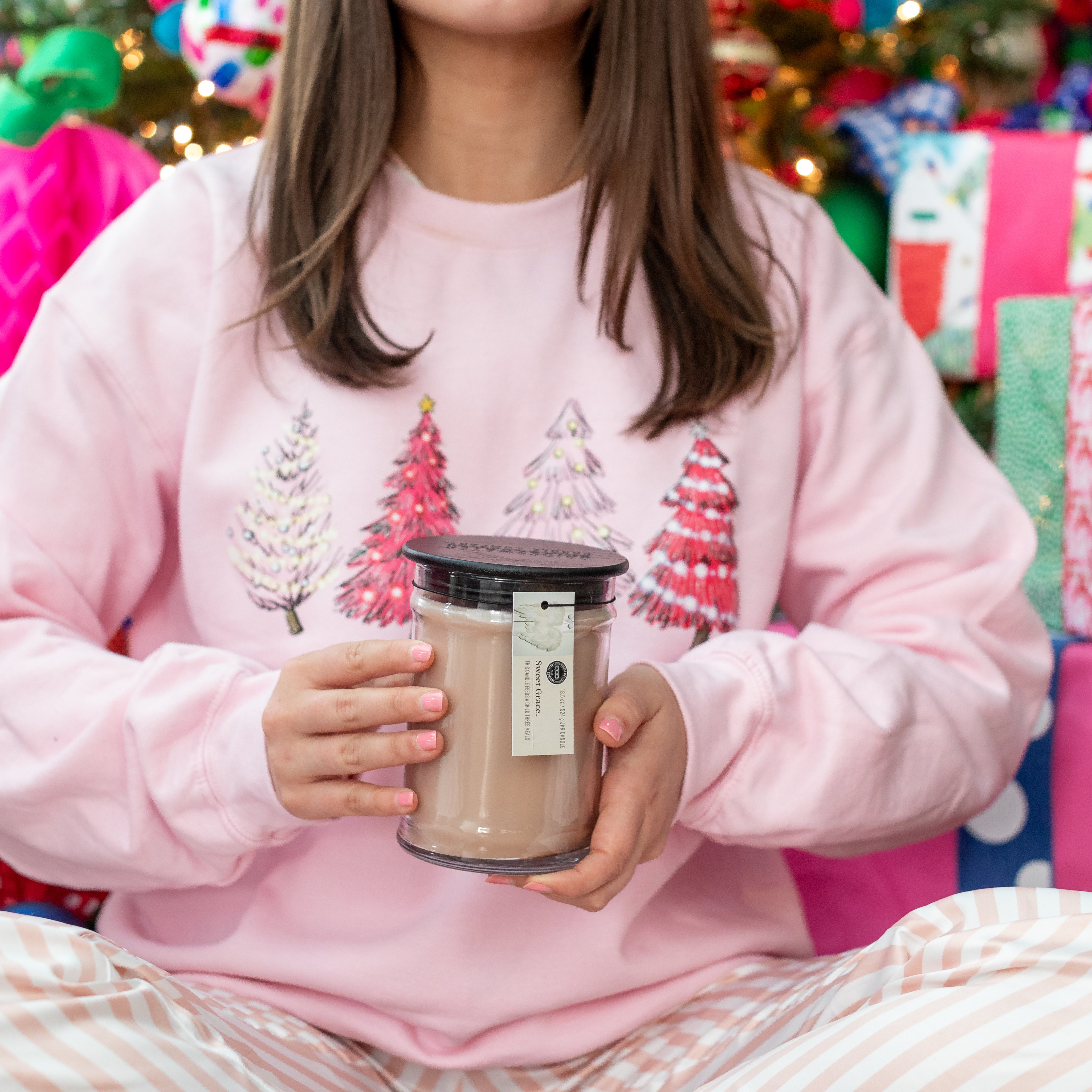 A person in pink and white pajamas and a pink Christmas tree sweatshirt holds a Bridgewater Candle Sweet Grace 18oz Large Jar Candle, with colorful gifts and a decorated tree in the background for festive fragrance charm.