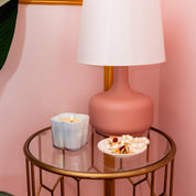 A round glass-top table holds a lit Bridgewater Candle Sweet Grace Collection Candle #077 in a blue holder, a white plate with pearl jewelry, and a pink lamp with a white shade. The background wall is pink.