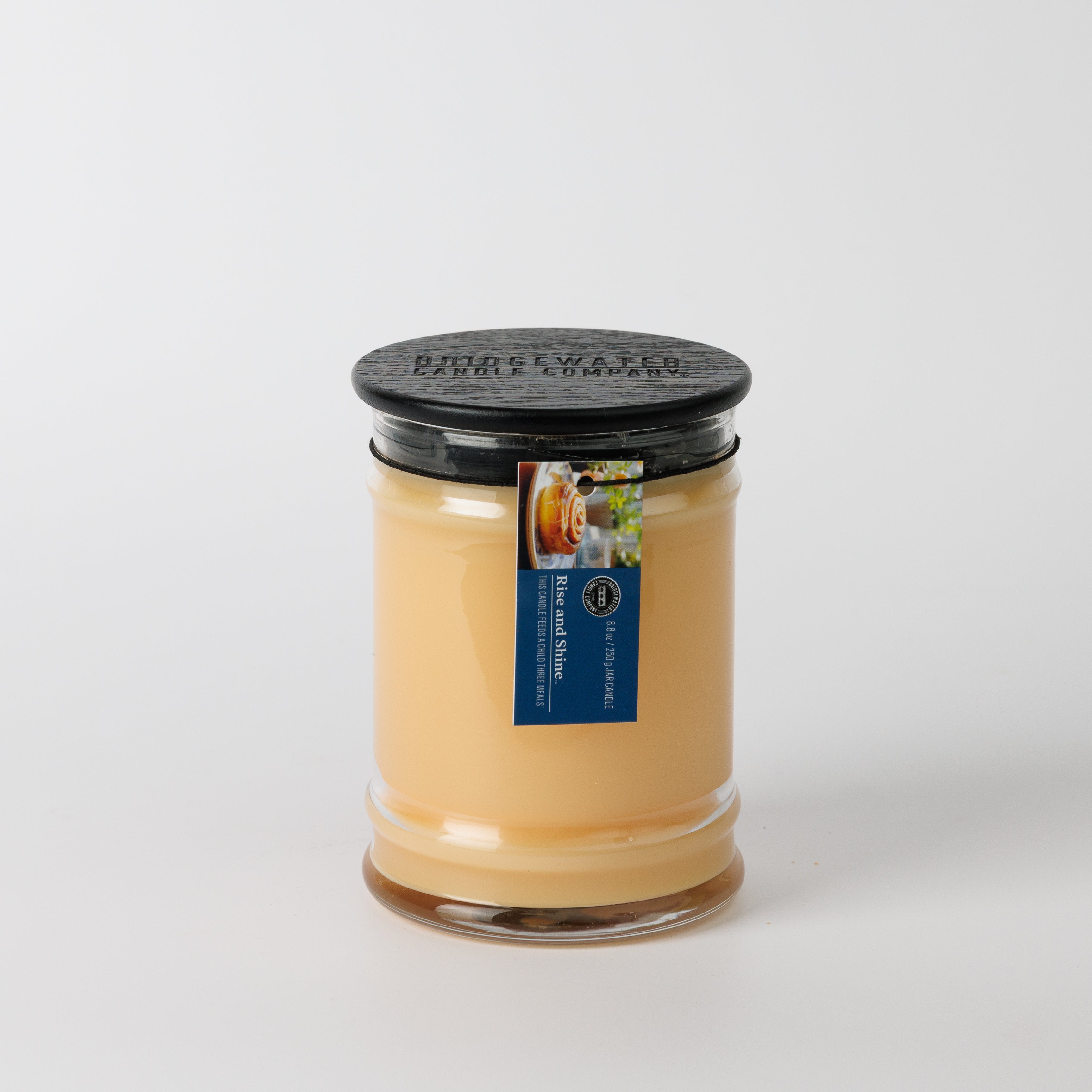 An 8oz glass jar candle by Bridgewater Candle, Rise & Shine, features soy-blend wax in light tan. Sealed with a black lid and adorned with a blue label showing pancakes, it sits against a plain white background.