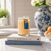A Bridgewater Candle Rise & Shine 18oz Large Jar Candle, crafted with soy-blend wax, glows in a glass jar atop two books next to a blue and white floral vase with white flowers, while a bowl of oranges sits on the marble countertop in sunlight.