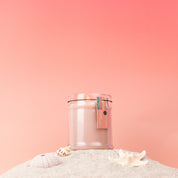 The Sunkissed 8oz Jar Candle by Bridgewater Candle, made with soy-blend wax, sits on sand amid seashells and a starfish against a soft pink gradient. A tag with blue and pink details hangs from the clear glass jar.